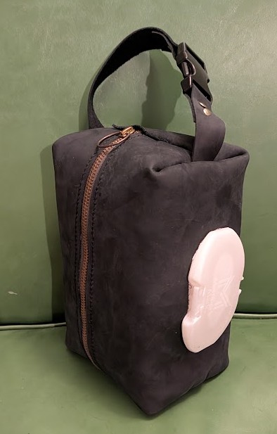 a blocky-shaped pouch made of black leather, with the lid of a baby-wipe container sticking out of one side. a strap on the pouch is secured with a buckle