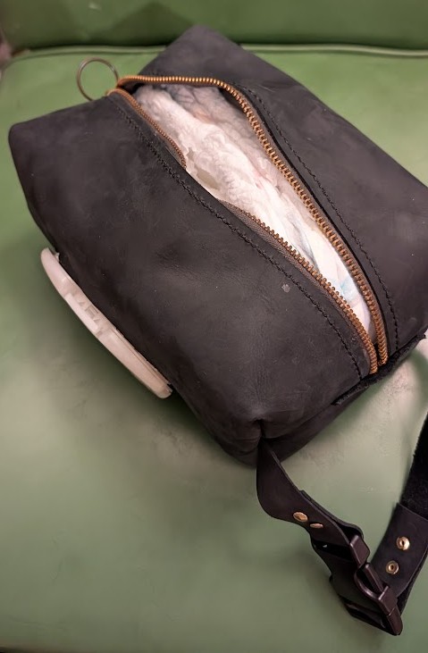 a blocky-shaped pouch made of black leather, with the lid of a baby-wipe container sticking out of one side. a strap on the pouch is secured with a buckle