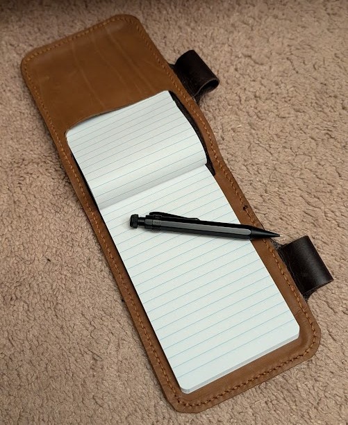 a dark brown leather notepad cover, with a black mechanical pencil hanging through a pen loop on one side